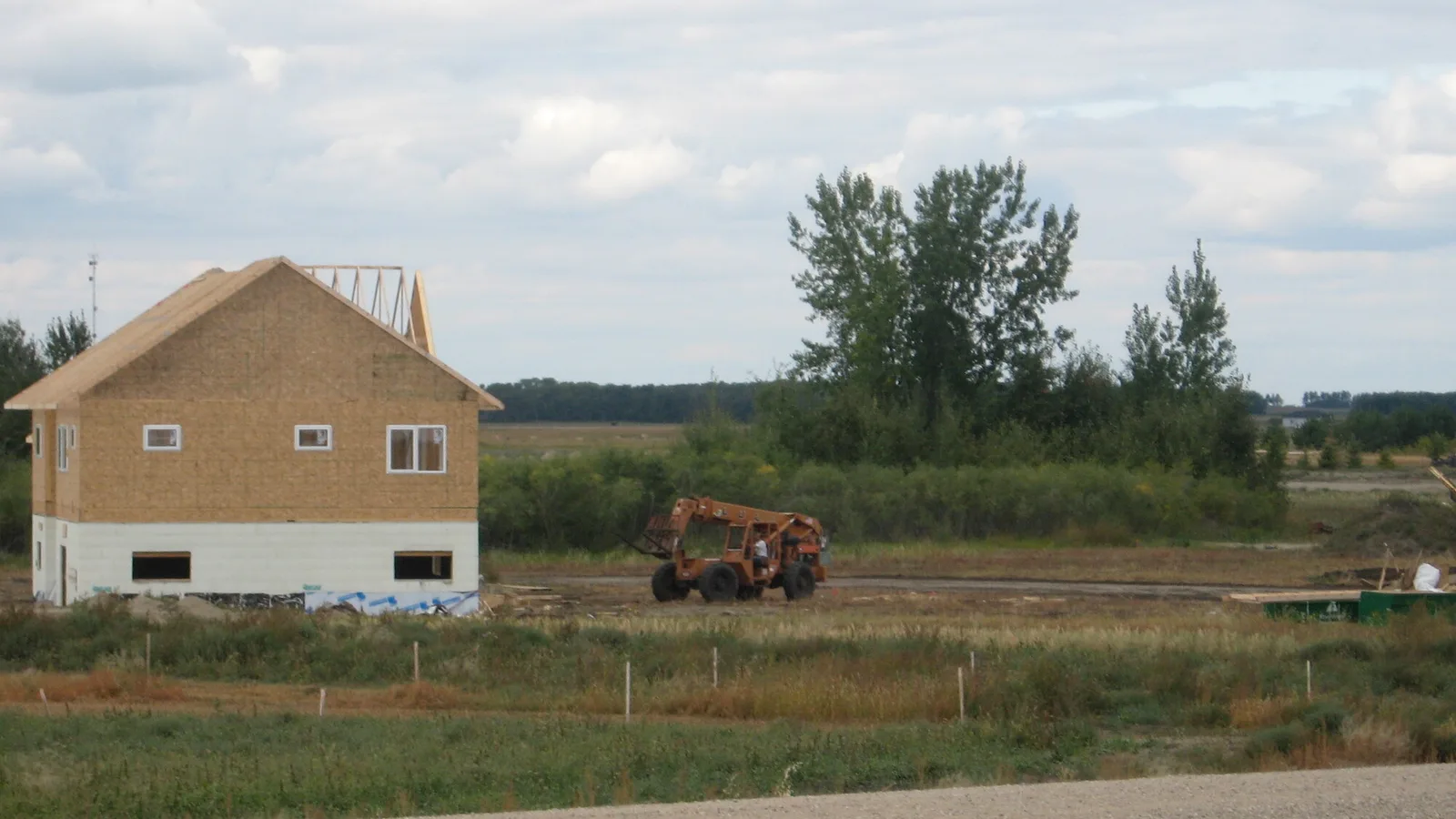 A house under construction with exposed wooden wall framing, roof trusses and a white insulated concrete form foundation, in a new housing development near Sask